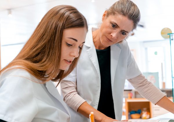 A female blonde pharmacist helps and assists her young aprrentice trainee during studies of medical books